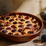 Clafoutis aux prunes dore et moelleux dans un plat en terre cuite, prunes violettes noyees dans la pate cremeuse, lumiere provencale chaude