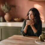 Femme sereine tenant une tasse de tisane dans une cuisine méditerranéenne lumineuse, symbole d'attention portée à son corps